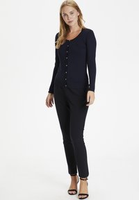 Jūras spēku garš piedurknes adīts kardigāns izgatavots no adītas auduma, ar apaļu kakla izgriezumu, pogu aizdari un ribotu apakšmalu. Saskaņots ar melnām šaurajām biksēm un augstpapēžu kurpēm.