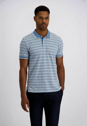 Mann trägt hellblaues Poloshirt mit weißen horizontalen Streifen und dunkle Hose, steht vor einem schlichten hellen Hintergrund.