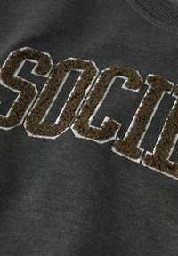 Tmavě šedá mikina s texturou, na které je fuzzy hnědý nápis "SOCIAL" orámovaný bílou barvou, s kulatým výstřihem a měkkým materiálem.