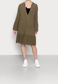 Olivgrön, lager-på-lager klänning med långa, utställda ärmar och V-ringning, bärs över en svart spets topp. Kombineras med vita sneakers.