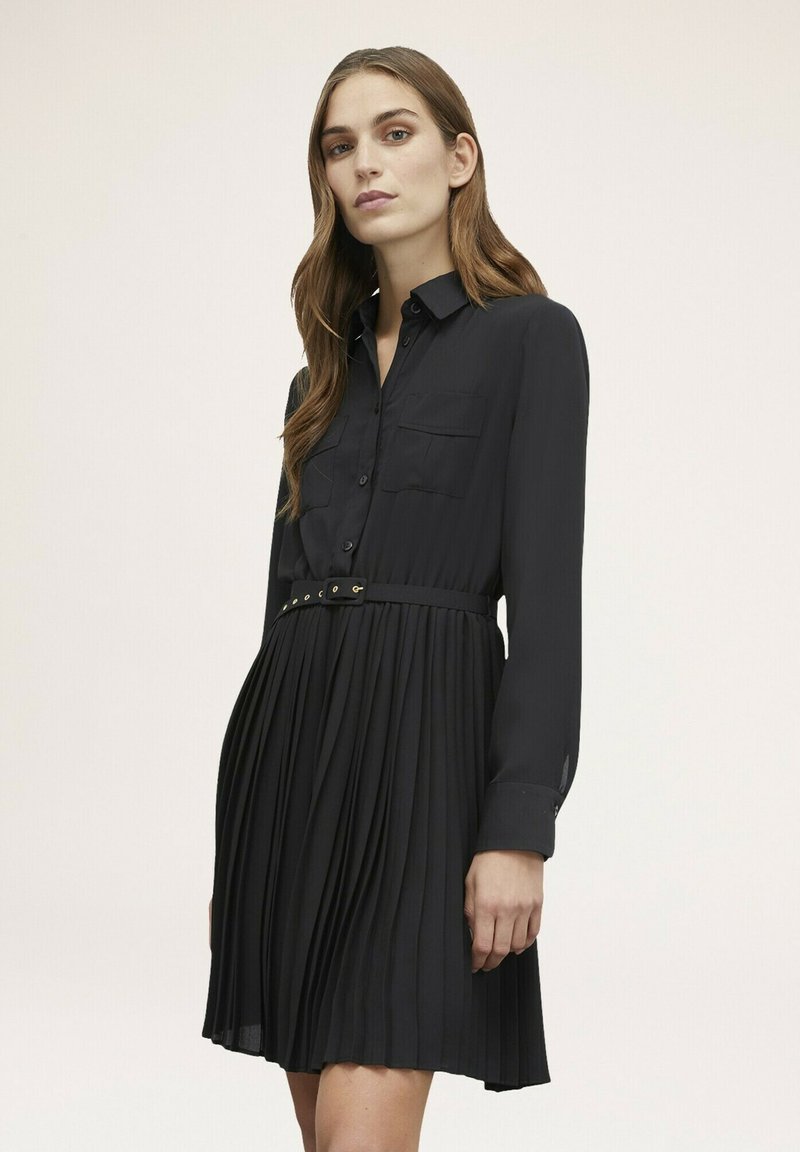 Abito camicia plissettato nero con maniche lunghe, chiusura a bottoni frontale, due tasche sul petto e vita con cintura. Tessuto liscio, lunghezza sopra il ginocchio.