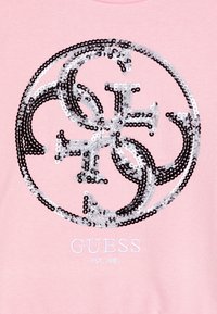 Sweat-shirt rose avec un logo circulaire composé de paillettes noires et argentées, et "GUESS" brodé en blanc en dessous du logo.