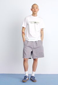 Witte t-shirt met groen grafisch logo, gecombineerd met lichtgrijze drawstring shorts. Blauwe sneakers met witte strepen en witte sokken.