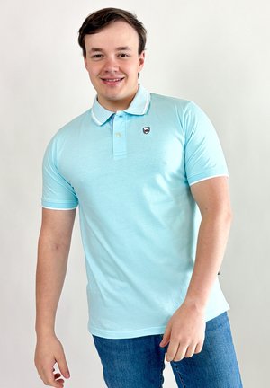 Jeune homme souriant, portant un polo bleu clair avec un petit emblème et un jean bleu, debout devant un fond uni clair.