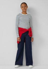 Een jongere staat tegen een grijze muur met een wit en marineblauw gestreept langemouwen shirt, brede spijkerbroek, witte schoenen en een rode sweater om de taille geknoopt.