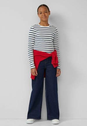 Een jongere staat tegen een grijze muur met een wit en marineblauw gestreept langemouwen shirt, brede spijkerbroek, witte schoenen en een rode sweater om de taille geknoopt.