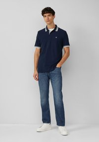 Marineblauwe polo met witte gestreepte kraag en manchetten, gecombineerd met blauwe jeans en witte sneakers, weergegeven tegen een effen achtergrond.