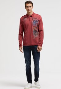 Langärmliges rotes Polohemd mit Grafikdruck, Knopfkragen und blauen Akzenten. Kombiniert mit dunklen Jeans und weißen Turnschuhen.