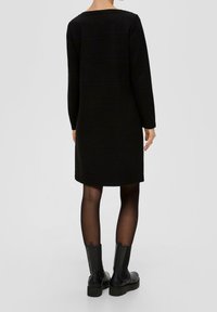 Robe noire à manches longues avec une coupe droite, en tissu texturé et col rond. Associée à des bottines noires et des collants transparents.