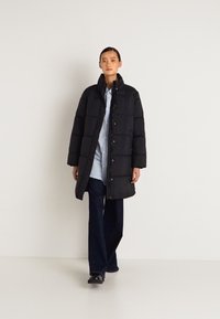 Ženska v dolgem črnem puhovem plašču, oblečena v modro srajco in temne hlače, hodi naprej proti enotni svetli ozadju.