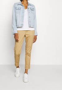 Veste en jean bleu clair avec un design légèrement usé, portée sur un t-shirt blanc à col en V, associée à un pantalon chino beige et des baskets blanches.