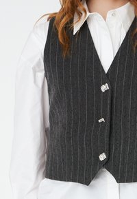 Gray pinstriped vest, tailored fit, featuring a v-neckline and three shiny silver buttons. Worn over a white dress shirt.