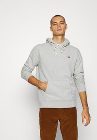 Lichtgrijze hoodie met trekkoord, voorzak en klein rood logo. Draagt met bruine corduroy broek, met lange mouwen en geribde manchetten.