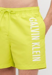 Shorts de bain vert lime en tissu lisse, présentant un logo "CALVIN KLEIN" blanc imprimé verticalement sur le côté gauche. Ceinture ajustable.