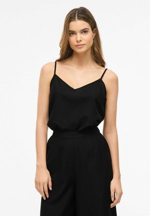 Kobieta z długimi brązowymi włosami, ubrana w czarny top bez rękawów z dekoltem w kształcie litery V oraz wysokim stanem, stojąca na białym tle.