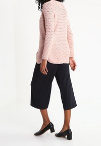 Suéter de punto rosa claro con un patrón de diamantes, combinado con culottes negros de pierna ancha y zapatos de tacón negros. Vista trasera.