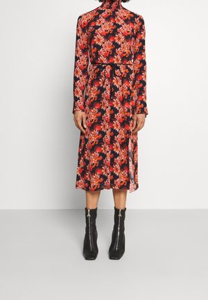 Femme portant une robe fleurie à manches longues et col montant avec des fleurs rouges et roses, et des bottines noires avec fermetures éclair à l’avant.