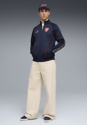 Jonge man staat met handen in zijn zakken, draagt een marineblauwe Puma-jas met rode accenten, crèmewitte broek met brede pijpen, een marineblauwe pet en blauwe schoenen.