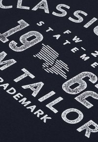 El tejido de color azul marino presenta grandes letras en blanco desgastadas. Las palabras incluyen "CLÁSICO", "SASTRE" y "1962", con acentos gráficos de rayas horizontales.