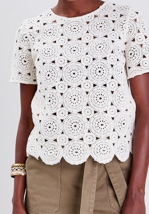 Biały krótki top na szydełku z okrągłymi wzorami, noszony z oliwkowozielonymi spodniami z wysokim stanem i złotymi bransoletkami na lewym nadgarstku.