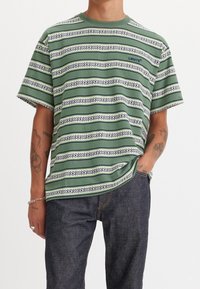 Mies, joka käyttää vihreää ja valkoista raidoitettua Levi's-teepparia, jossa on lyhyet hihat, ja tummansiniset farkut, yksi käsi taskussa.