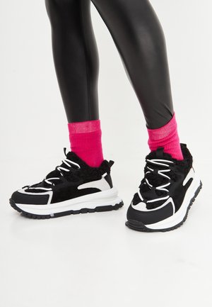 Jambes en leggings noirs brillants, chaussettes rose vif et baskets épaisses noires et blanches sur fond blanc.