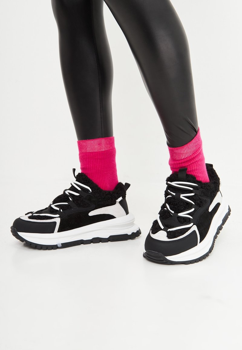 Jambes en leggings noirs brillants, chaussettes rose vif et baskets épaisses noires et blanches sur fond blanc.