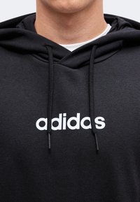 Άνδρας που φοράει μαύρο φούτερ Adidas με λευκό λογότυπο και κορδόνια, εμφανίζοντας τον λαιμό και το κάτω μέρος του προσώπου.