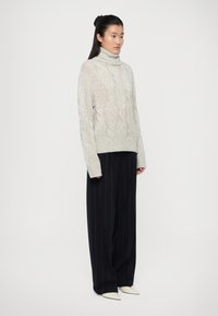 Pull à col roulé en maille torsadée gris clair avec ourlet côtelé, associé à un pantalon large à rayures noires et des chaussures blanches à bout pointu.