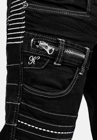 Pantaloni in denim neri con cuciture bianche, tasca con zip e design ricamato a "K". Vestibilità slim con dettagli a pannello sui lati.