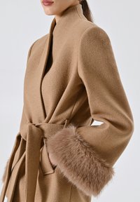 Cappotto avvolgente beige in lana con colletto alto, cintura e morbidi polsini in pelliccia sintetica. Presenta un tessuto testurizzato e due tasche laterali.