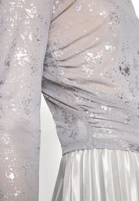 Silber- und graues Kleid mit einem durchsichtigen, glitzernden Oberteil mit gerafften Seiten und langen Ärmeln, kombiniert mit einem plissierten Satinrock.