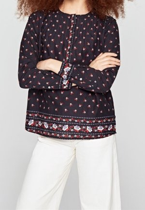 Femme portant un chemisier noir à manches longues avec de petits motifs floraux rouges et blancs et un pantalon blanc à jambes larges, bras croisés.
