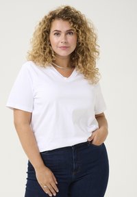 Camiseta blanca de manga corta y escote en V hecha de algodón, combinada con jeans de mezclilla azul oscuro. Cabello rizado y un sencillo collar de plata.