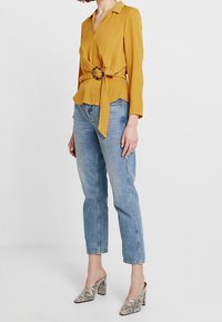 Donna che indossa una blusa gialla senape con cintura, jeans azzurri a gamba dritta e scarpe con tacco a blocco a motivo serpente, in piedi su un pavimento bianco.