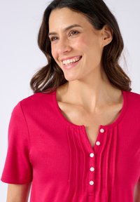 Top corto de color fucsia con mangas cortas y escote redondo, que presenta pliegues verticales y una tapeta de botones. Confeccionado con una tela suave y elástica.