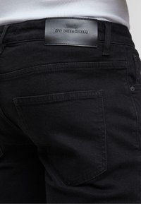 Gros plan d'une personne portant un jean noir avec une étiquette en cuir portant la mention "ZV PREMIUM" sur la ceinture et une poche arrière visible.