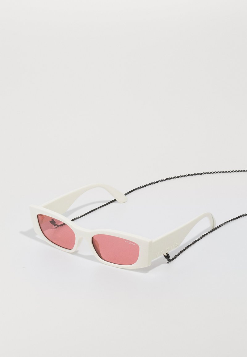 Hvit solbriller med rektangulære rosa linser, og en matte finish, samt en svart kjede for å bære rundt halsen. "Vogue"-logoen vises.