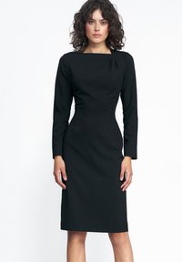 Czarna sukienka z długimi rękawami, o kwadratowym dekolcie, dopasowanym torsie i spódnicy do kolan. Gładki materiał z delikatnym marszczeniem przy ramieniu.