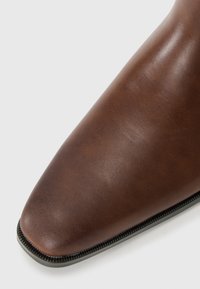 Bota de cuero marrón hasta el tobillo con punta afilada, textura suave y un detalle de cremallera negra contrastante a lo largo del costado.