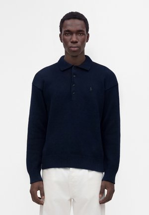 Homme portant un pull polo à manches longues bleu marine foncé et un pantalon blanc, debout devant un fond blanc uni.