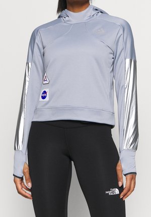 Person trägt einen hellgrauen Hoodie mit silbernen Ärmeln und NASA-Aufnähern, schwarze Leggings mit The North Face-Logo.
