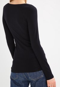 Schwarzes, langärmliges, enganliegendes Shirt mit rundem Ausschnitt. Glatte Textur, minimale Nähte, kombiniert mit blauen Denim-Jeans im klassischen Schnitt.