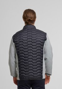 Veste hybride pour homme avec un devant matelassé noir texturé et des manches en tricot gris, dotée d'un col montant et de poignets élastiques.