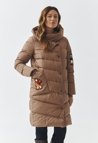 Gewatteerde bruine jas met hoge kraag, voorzien van decoratieve teddybeer patches op de zakken en drukknoopsluitingen. Soepele textuur, knielang.