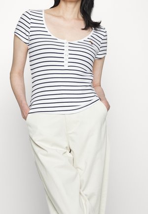 Camiseta de manga corta y cuello redondo en blanco con rayas azul marino, presenta un tapón de botones y un pequeño acento bordado. Lleva puesta con pantalones color crema.