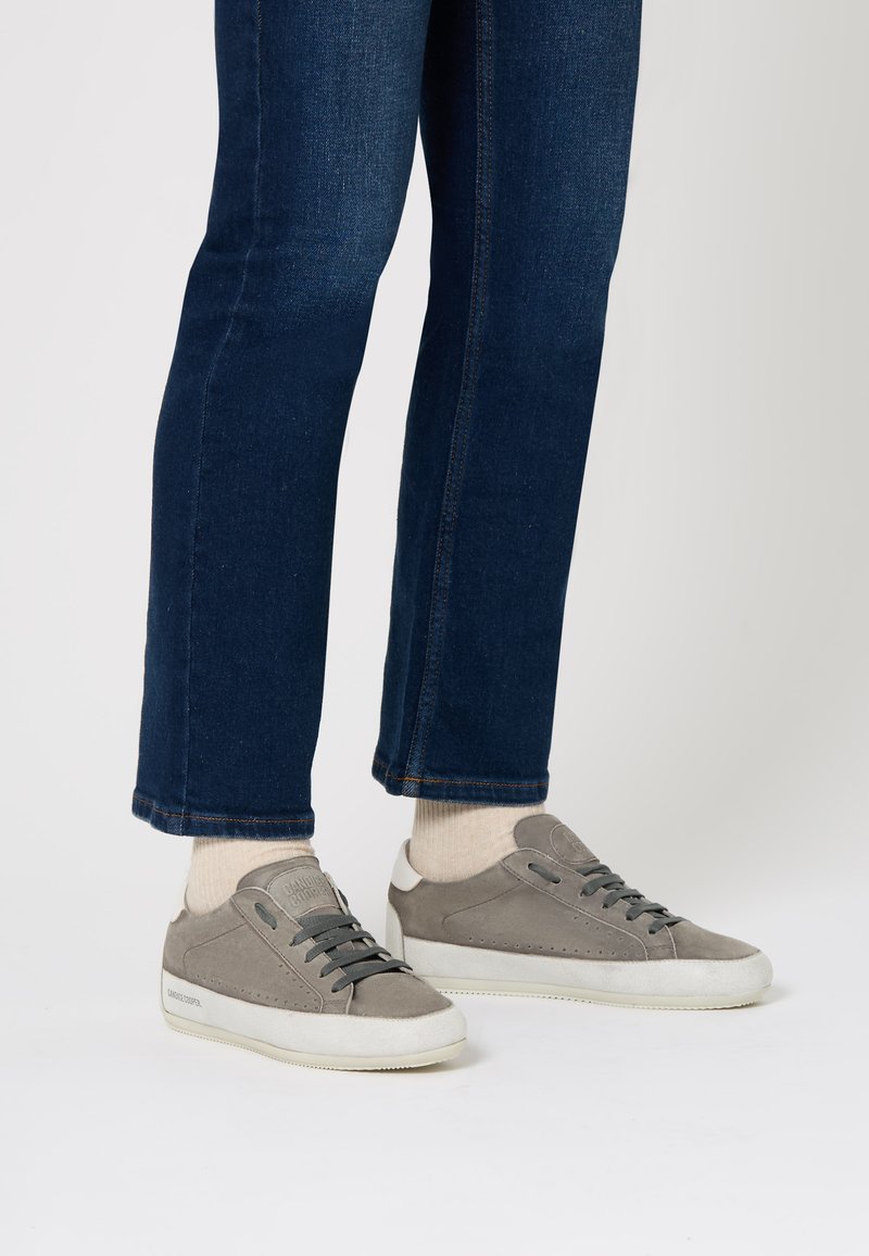 Sneakers in suede grigio con suola in gomma bianca, dotati di lacci e dettagli perforati. Abbinati a jeans blu scuro e calzini di colore chiaro.