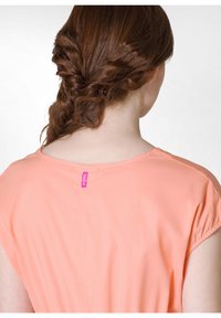 Persikofärgad skjorta med korta ärmar, med rundad halsringning och en liten rosa etikett bak. Mjuk tyg, slät yta.