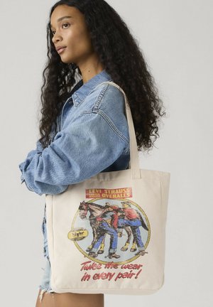 Nuori nainen kiharat hiukset, denimtakki päällä ja kantaa beigeä kangaskassia, jossa on vintage Levi's -kuvio ja teksti.