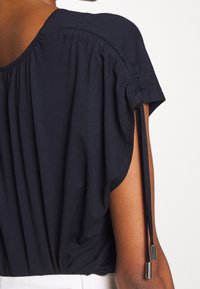 Námořnická modrá blůza s nařaseným materiálem na ramenou, krátkými rukávy a detailem se stahovací šňůrkou s kovovými konci. Hladký, jemný povrch.